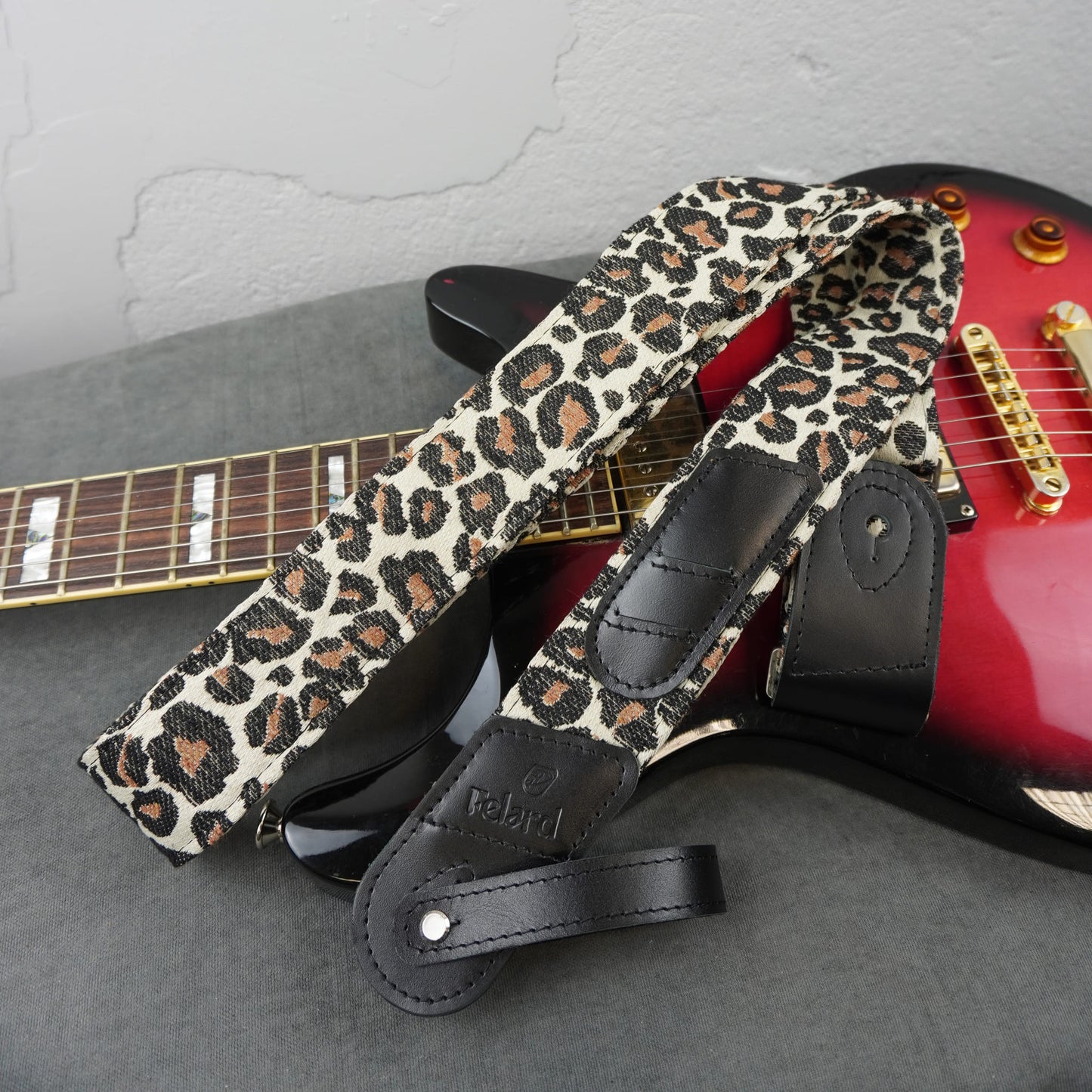 A leopard print guitar strap with black leather accents and a debossed 'Rebird' logo, draped over a red electric guitar on a gray surface against a light gray wall.