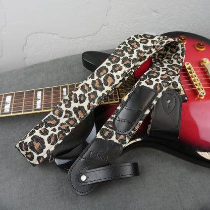 A leopard print guitar strap with black leather accents and a debossed 'Rebird' logo, draped over a red electric guitar on a gray surface against a light gray wall.
