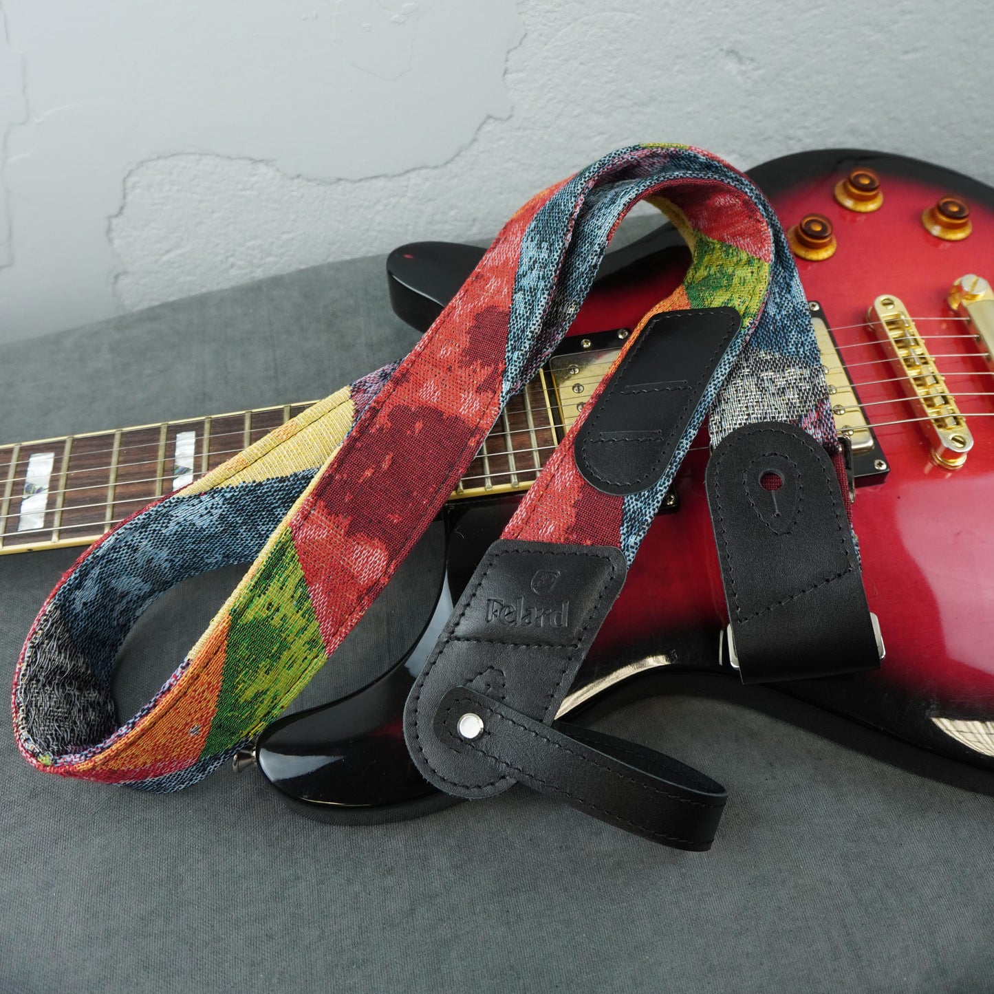 A colorful guitar strap with a multicolor abstract pattern and black leather end pieces, draped over a red electric guitar on a gray surface.
