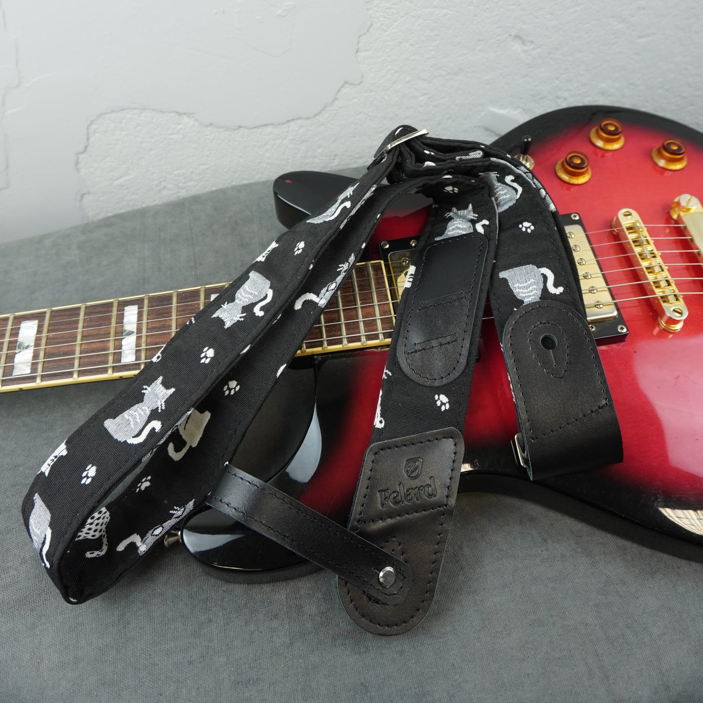 A black guitar strap with a white cat and paw print pattern, draped over a red electric guitar on a gray surface against a light gray wall.