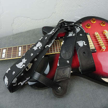 A black guitar strap with a white cat and paw print pattern, draped over a red electric guitar on a gray surface against a light gray wall.