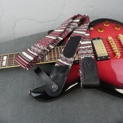 A guitar strap with a red, white, and black geometric pattern and black leather accents, draped over a red electric guitar on a gray surface.