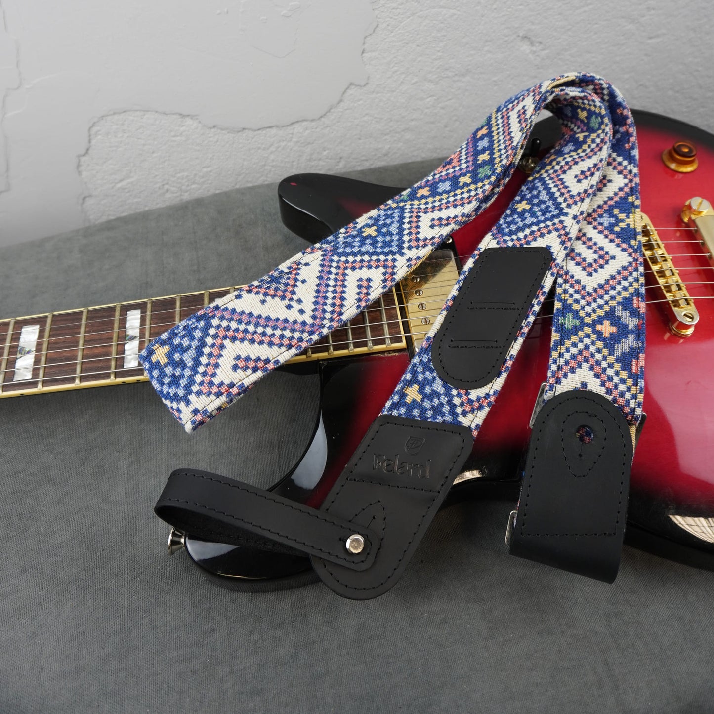 A guitar strap with a blue, white, and red geometric pattern and black leather ends, draped over a red electric guitar on a gray surface.