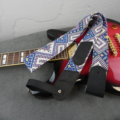 A guitar strap with a blue, white, and red geometric pattern and black leather ends, draped over a red electric guitar on a gray surface.