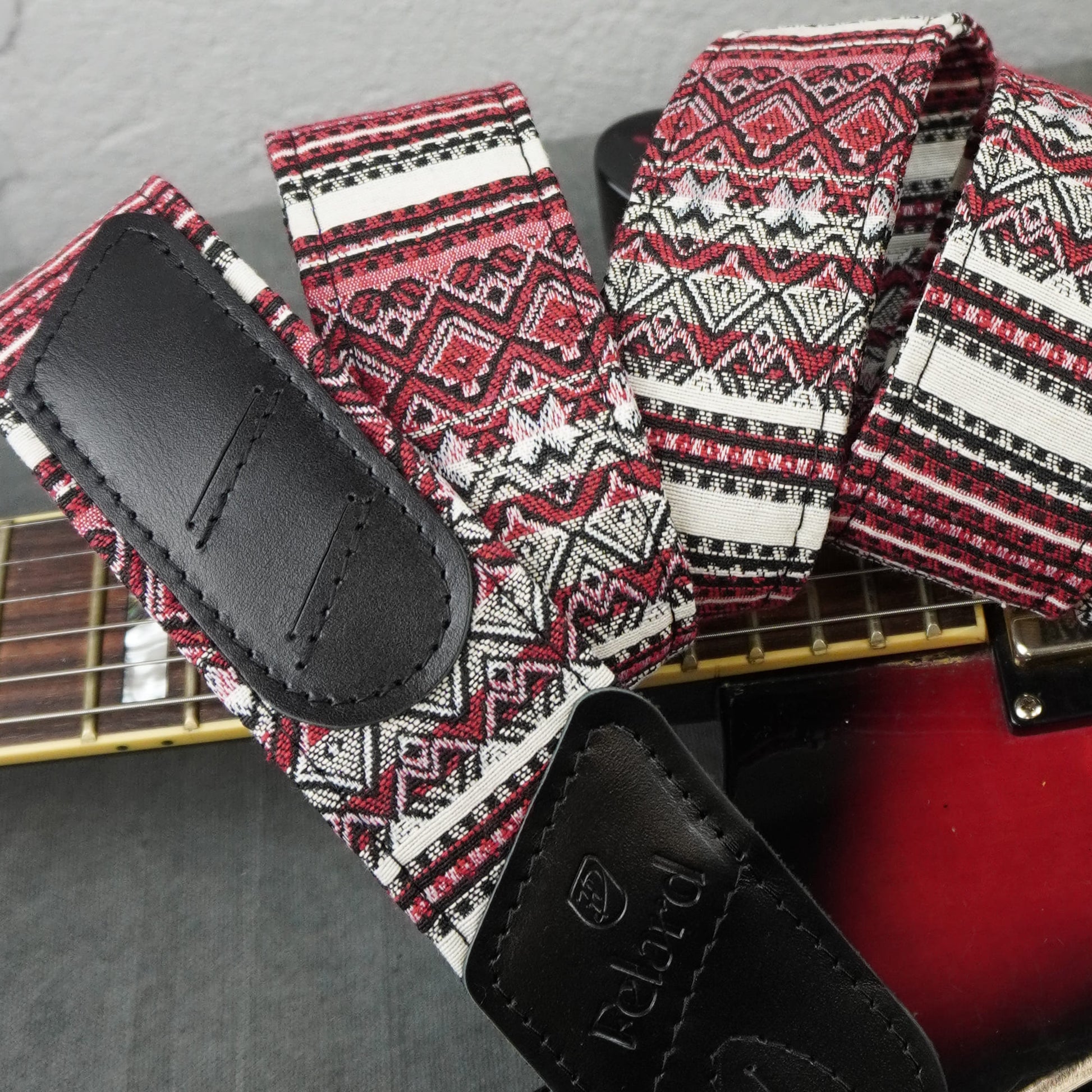 guitar strap with a red, black, and white geometric pattern and black leather ends, draped over the neck of a red electric guitar.