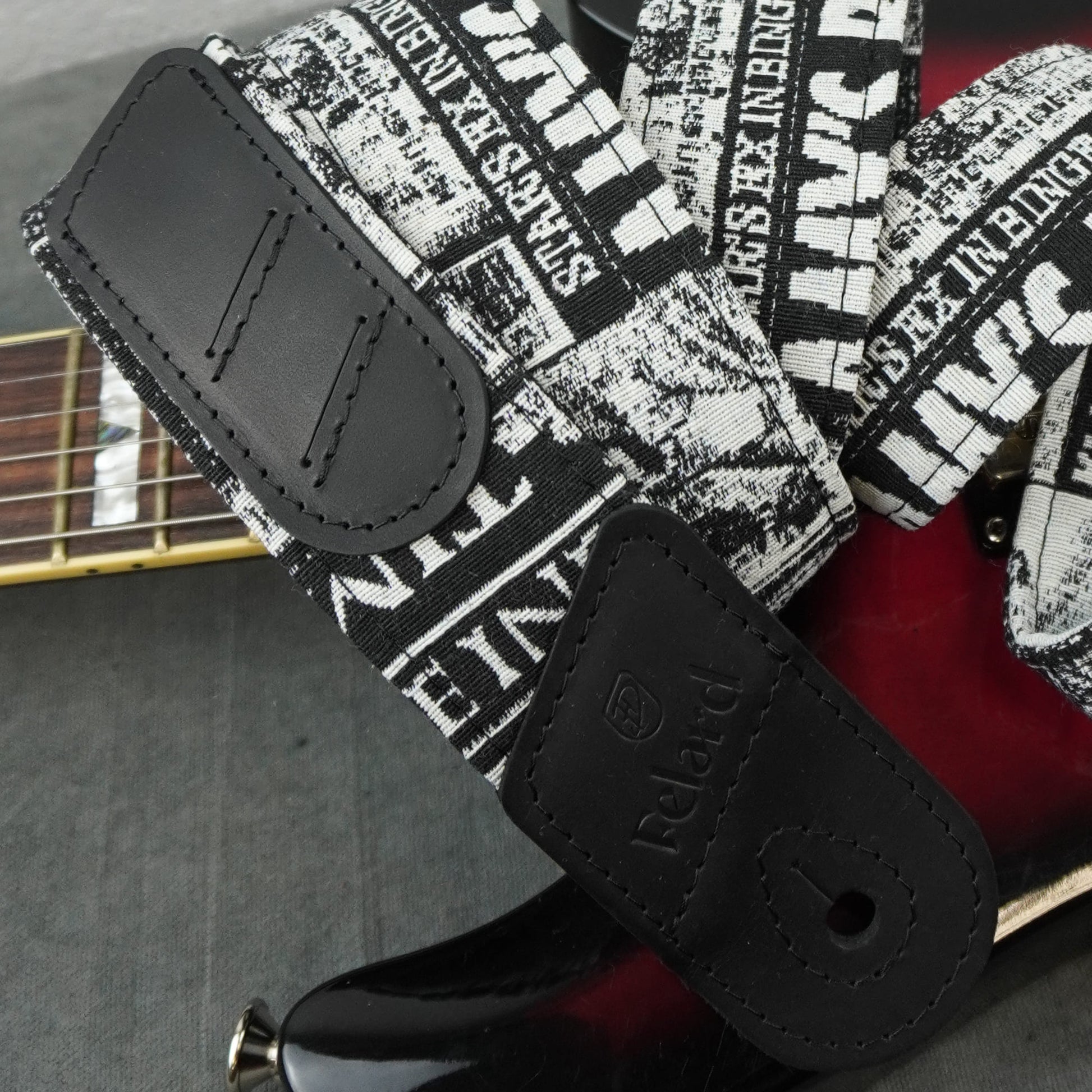 Felard electric guitar strap with a black and white newspaper print pattern and black leather end pieces, shown attached to a red electric guitar.