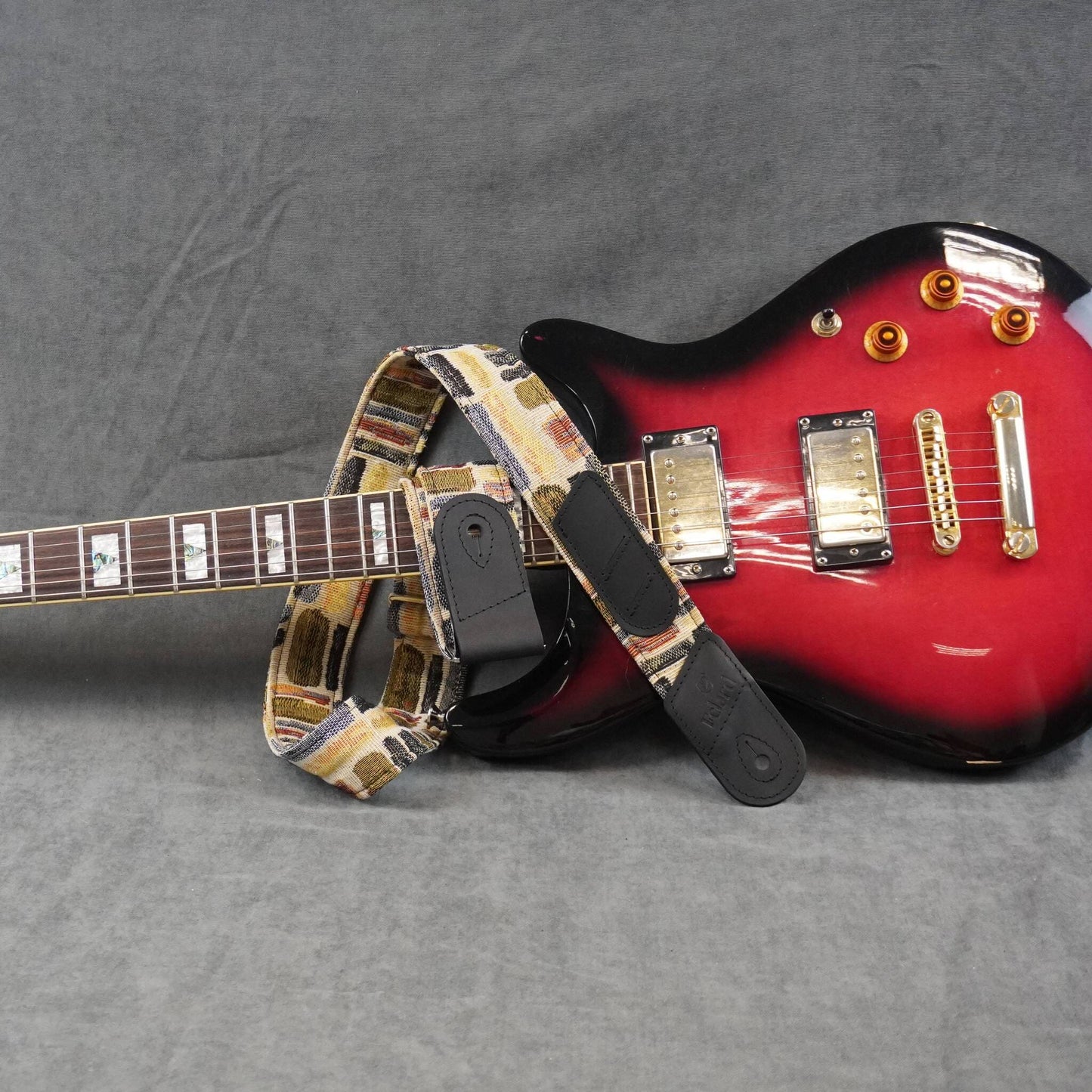 A guitar strap with a vintage cassette tape pattern in beige, brown, and black, shown draped over the neck and body of a red electric guitar on a gray textured background.