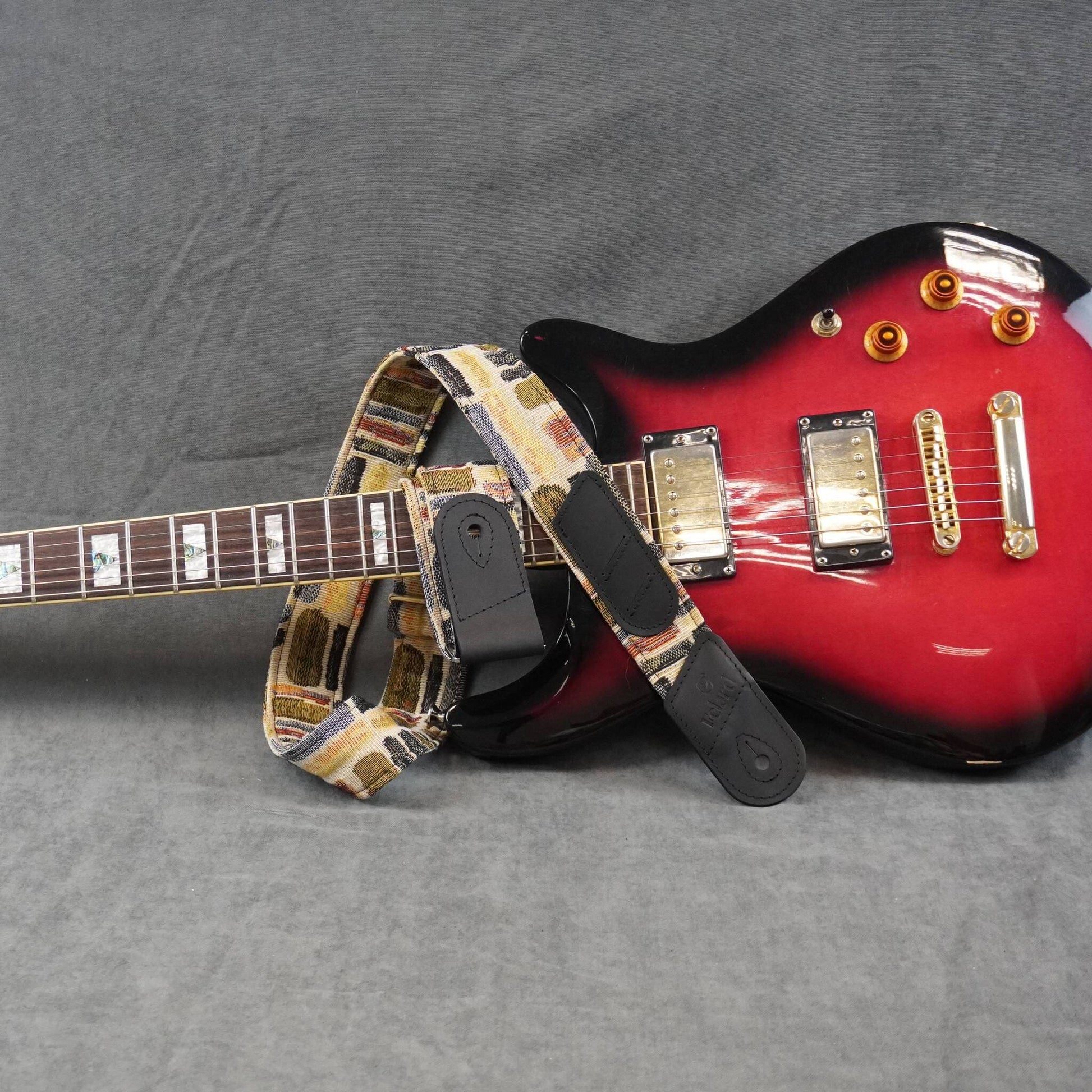A guitar strap with a vintage cassette tape pattern in beige, brown, and black, shown draped over the neck and body of a red electric guitar on a gray textured background.