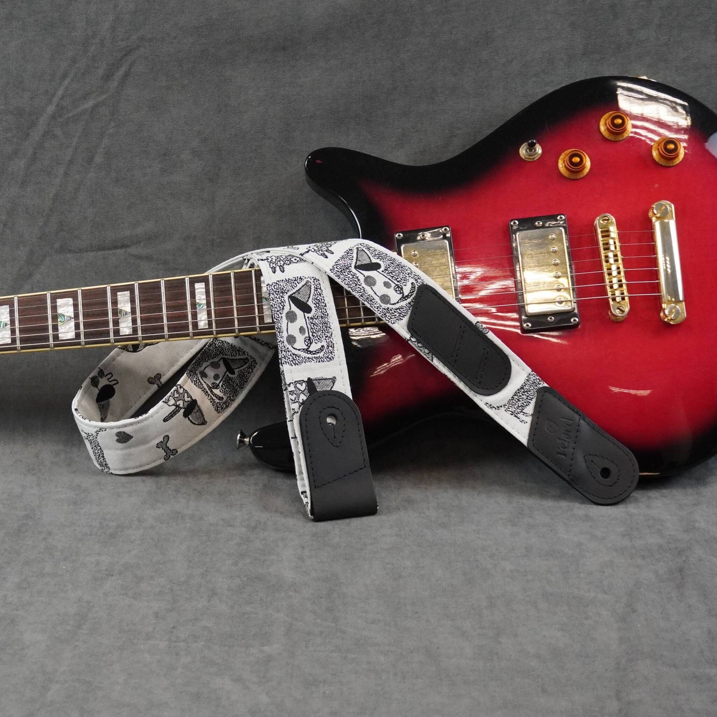 A guitar strap with a black and white skull and crossbones pattern, displayed on a red electric guitar against a gray fabric background.