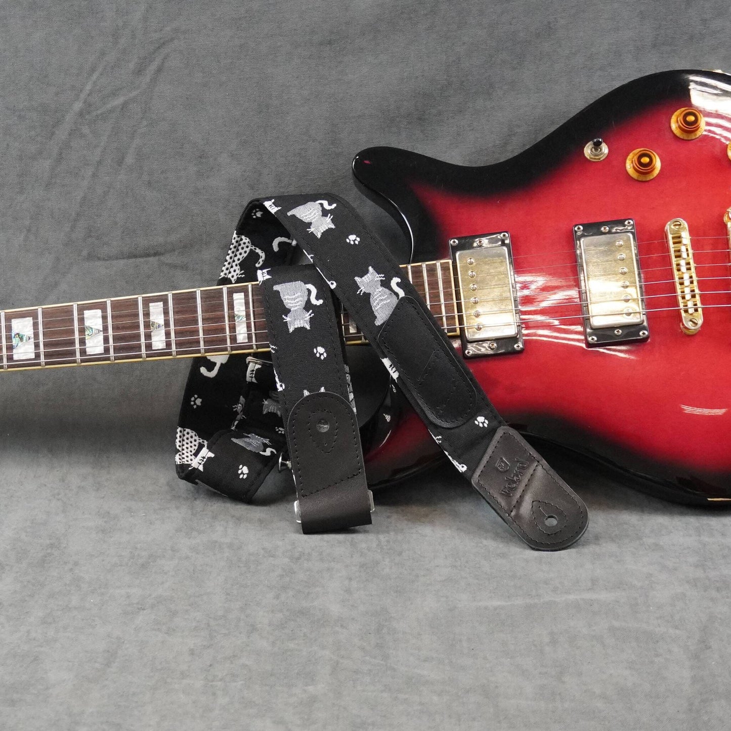 A black electric guitar strap with a white cat pattern, draped over a red and black electric guitar on a gray textured background.