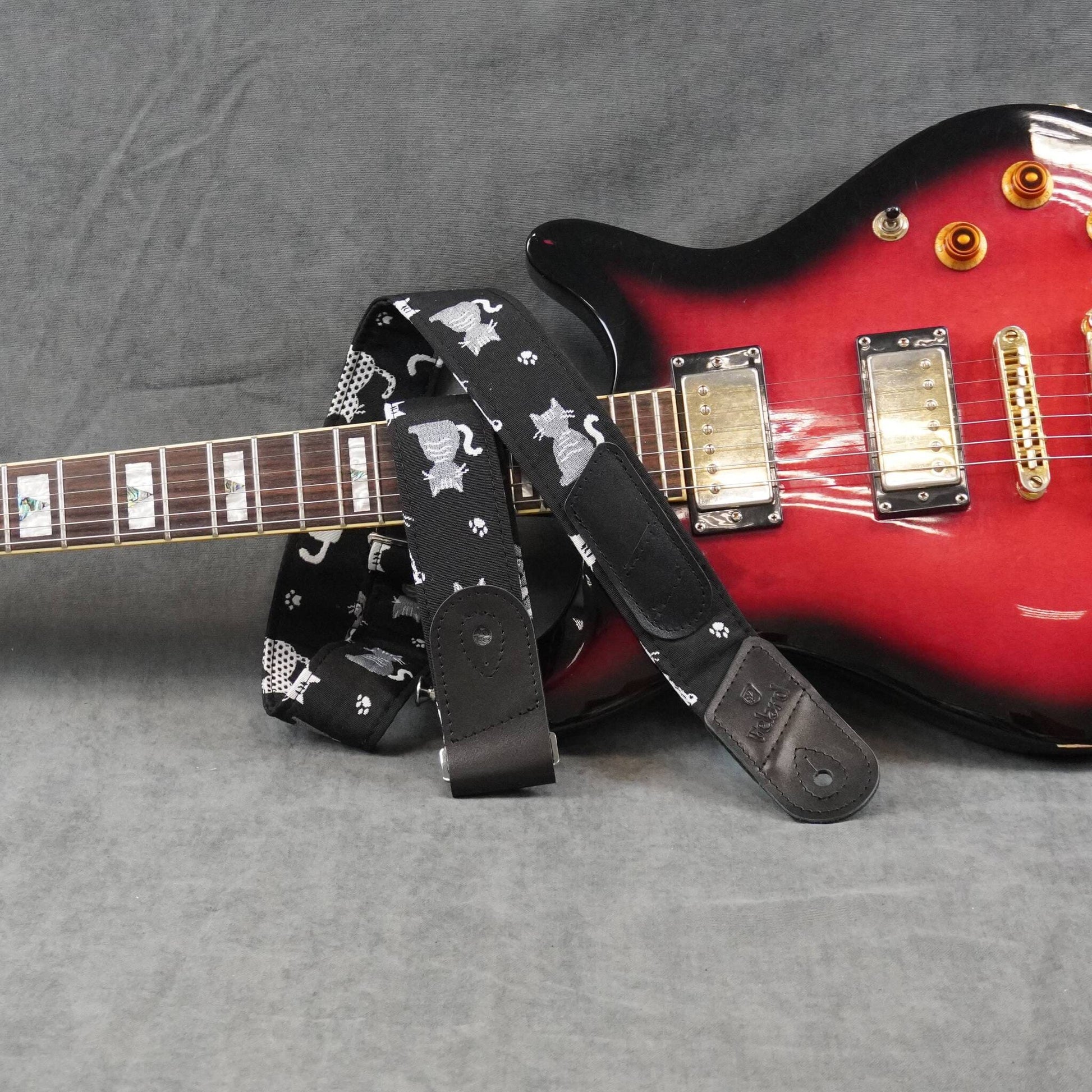 A black electric guitar strap with a white cat pattern, draped over a red and black electric guitar on a gray textured background.