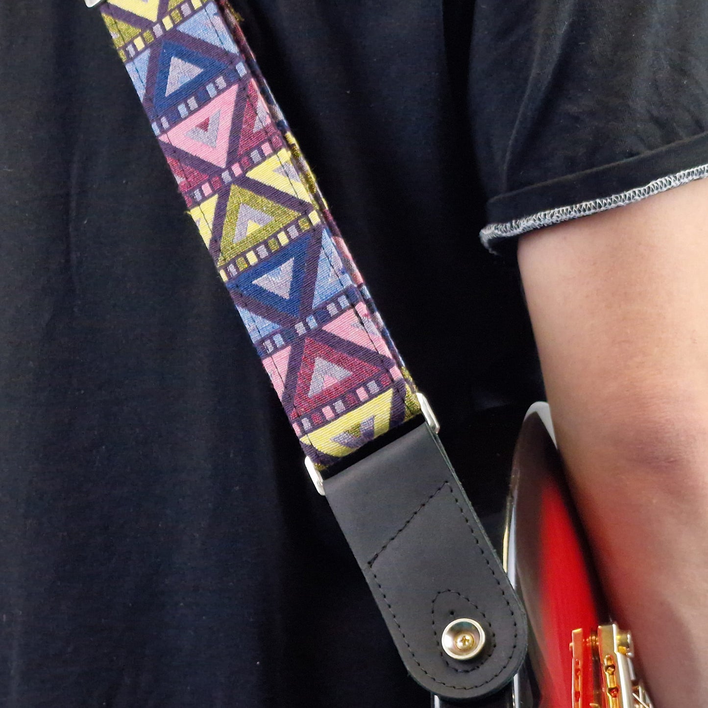 A close-up of a guitar strap worn across a person's torso. The strap has a colorful geometric pattern with pink, blue, yellow, and purple triangles, attached to a black leather end piece with a gold snap button.