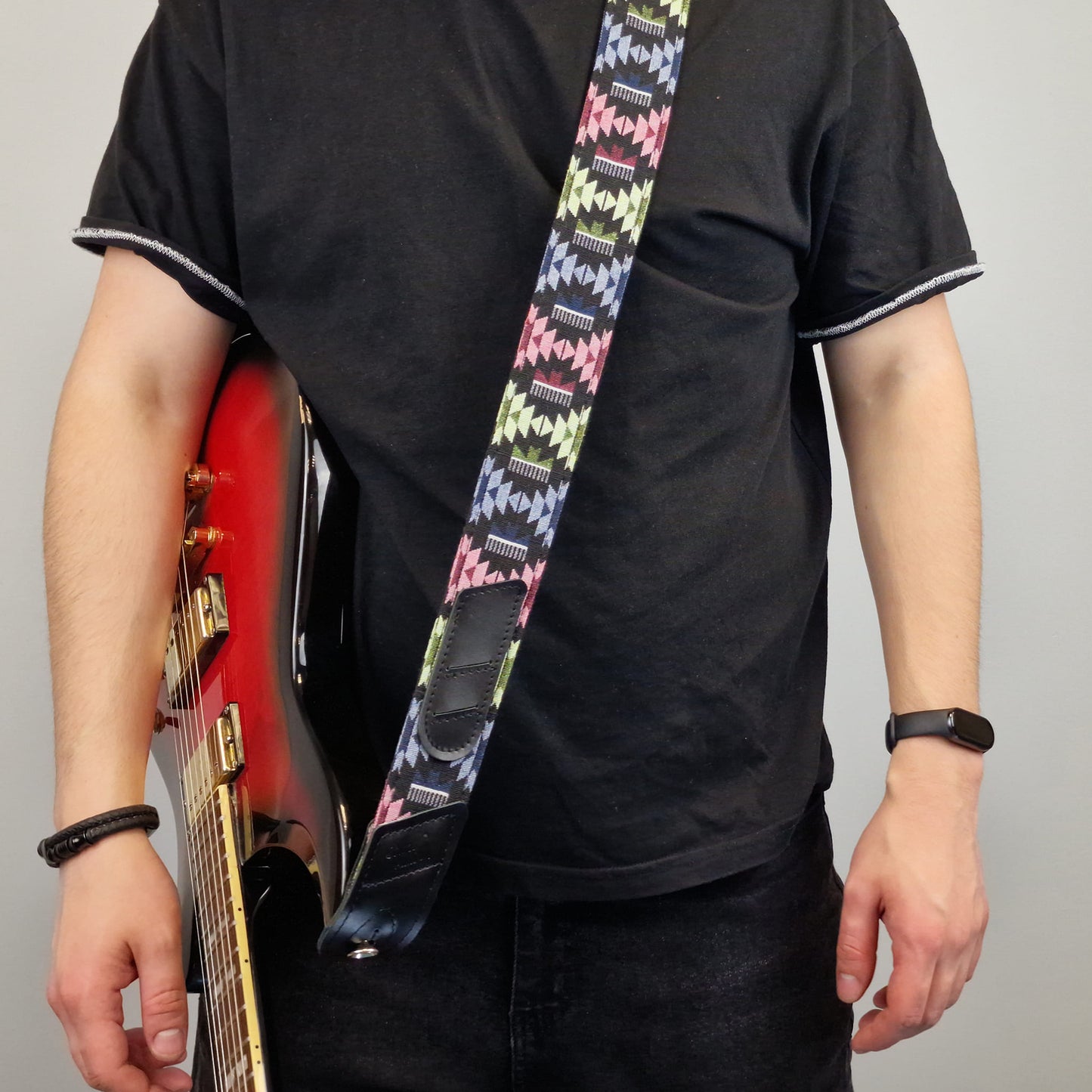  a colorful patterned guitar strap with a black central section. The strap features a repeating geometric design in pink, green, yellow, and blue on a black background.