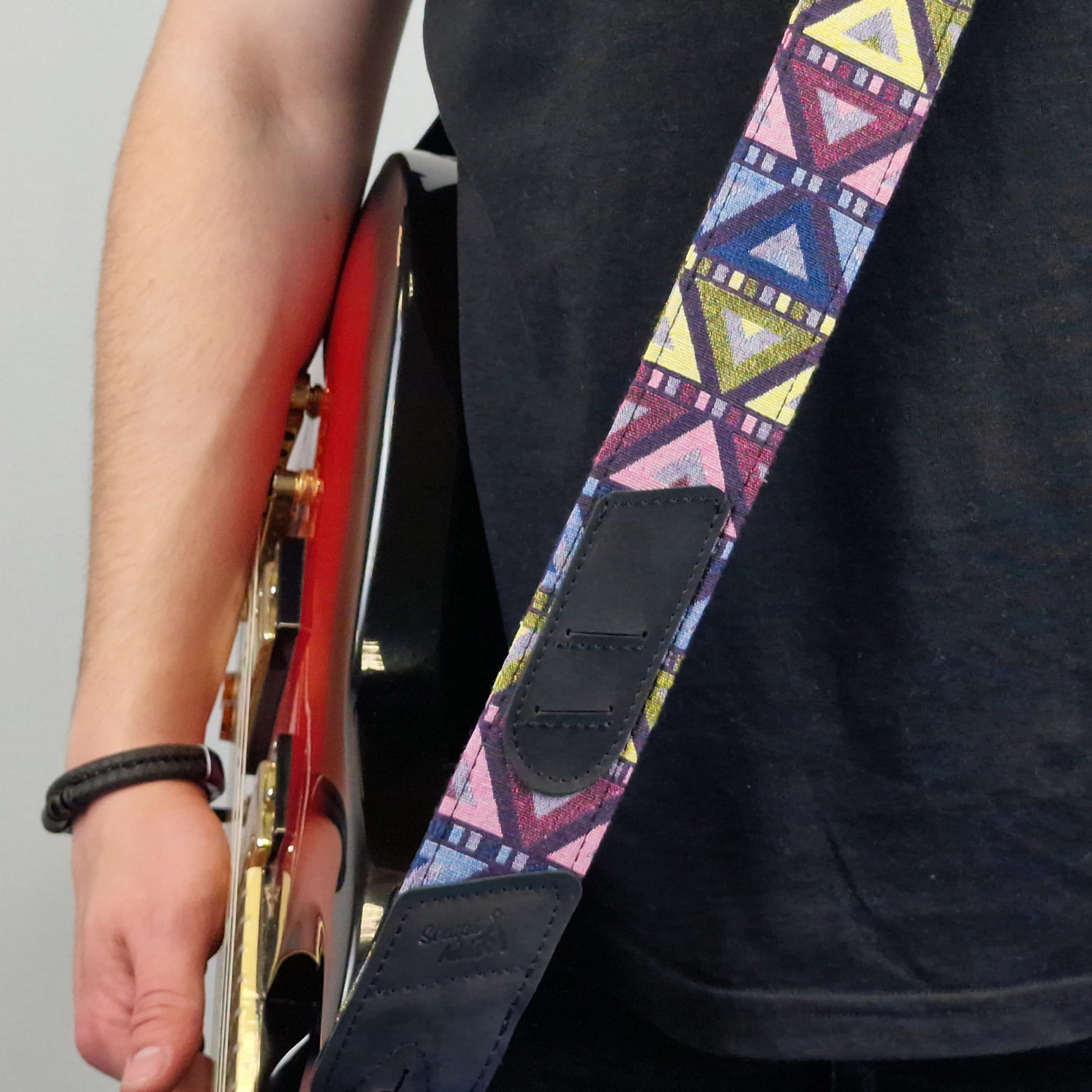 A close-up of a guitar strap with a colorful geometric pattern in pink, blue, yellow, and purple, featuring dark brown leather patches. The strap is worn by a person holding a red electric guitar.