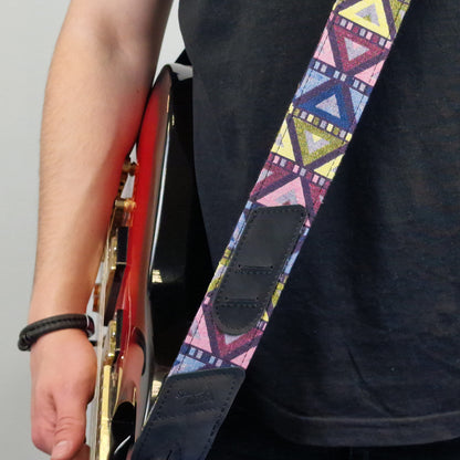 A close-up of a guitar strap with a colorful geometric pattern in pink, blue, yellow, and purple, featuring dark brown leather patches. The strap is worn by a person holding a red electric guitar.