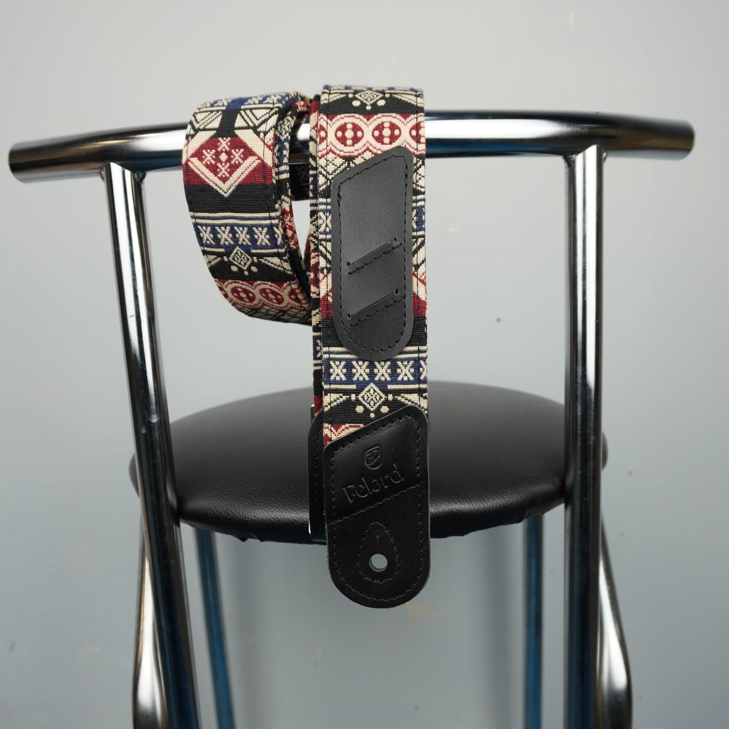 guitar strap with a colorful geometric pattern and black leather hardware, draped over the back of a black chair with a chrome frame.