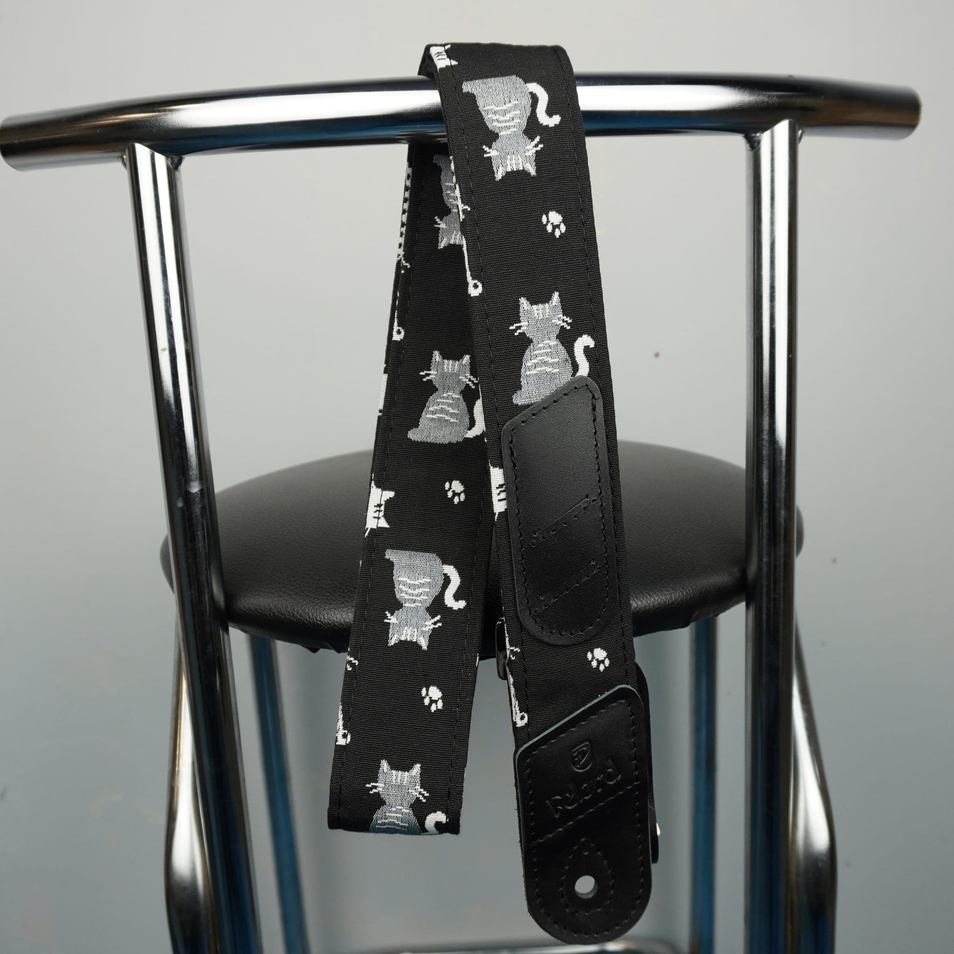 A black guitar strap with a repeating pattern of gray cats and paw prints, draped over the back of a chrome metal stool.