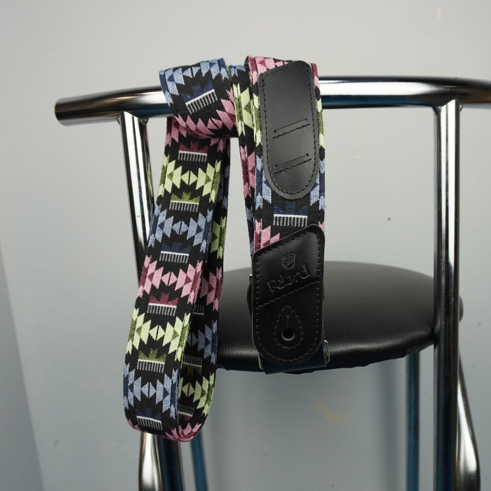 Felard guitar strap with a colorful geometric pattern and black leather ends, hanging from the back of a metal chair.