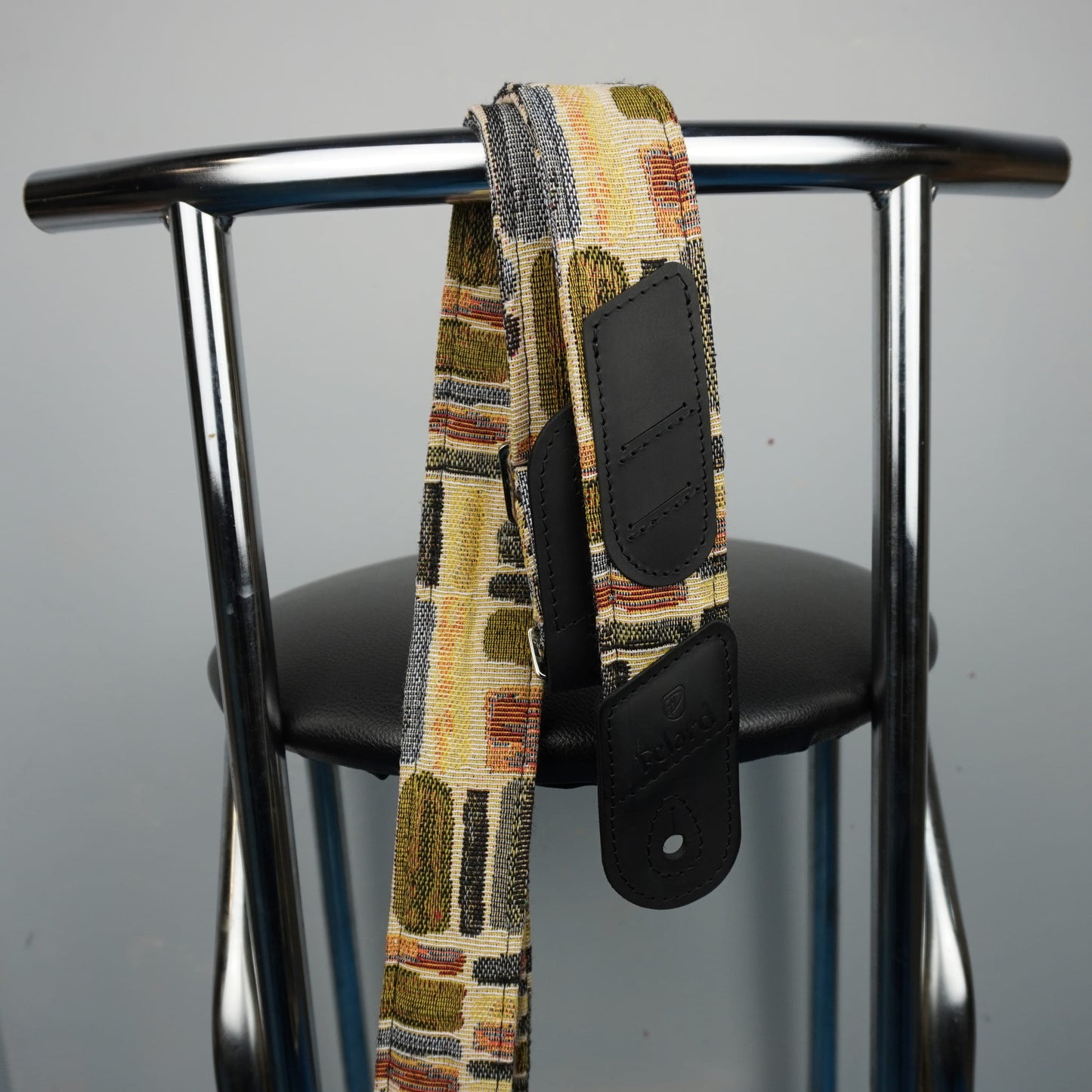 guitar strap with black leather hardware, draped over the back of a black chair with a chrome metal frame.
