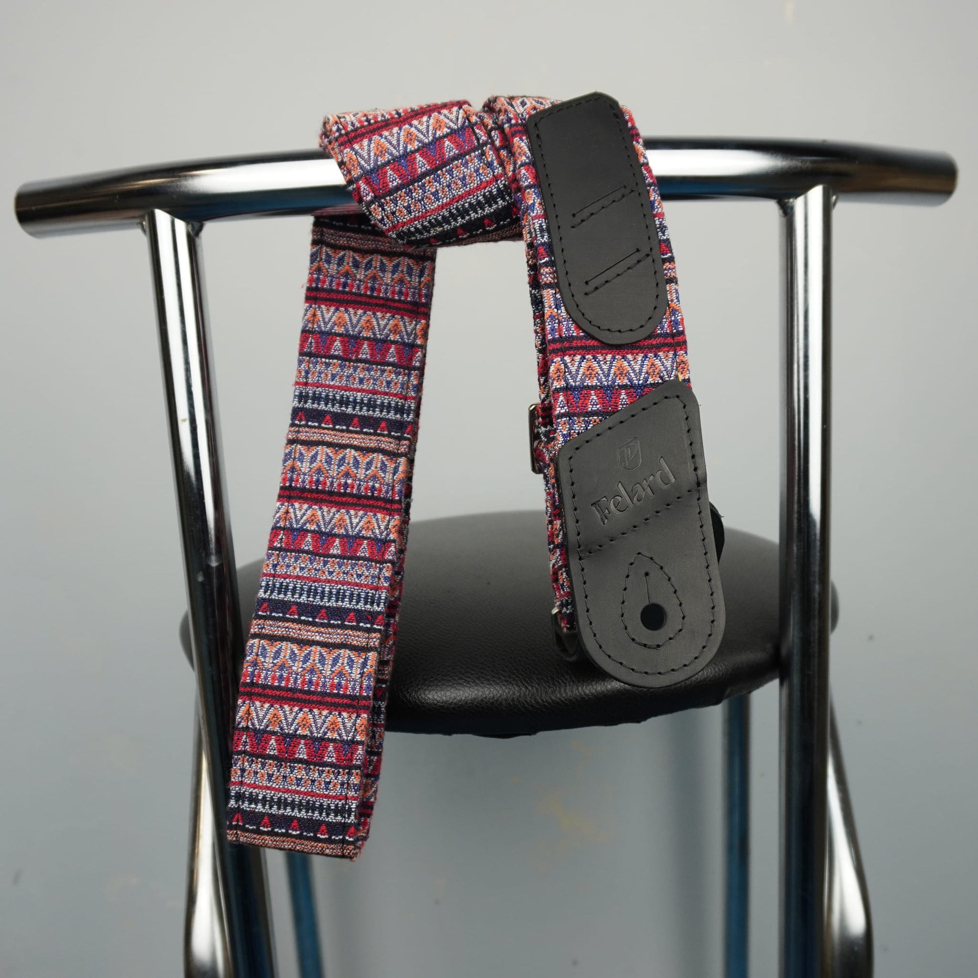A guitar strap with a red, blue, and white geometric pattern, draped over the back of a black chair with a chrome metal frame.