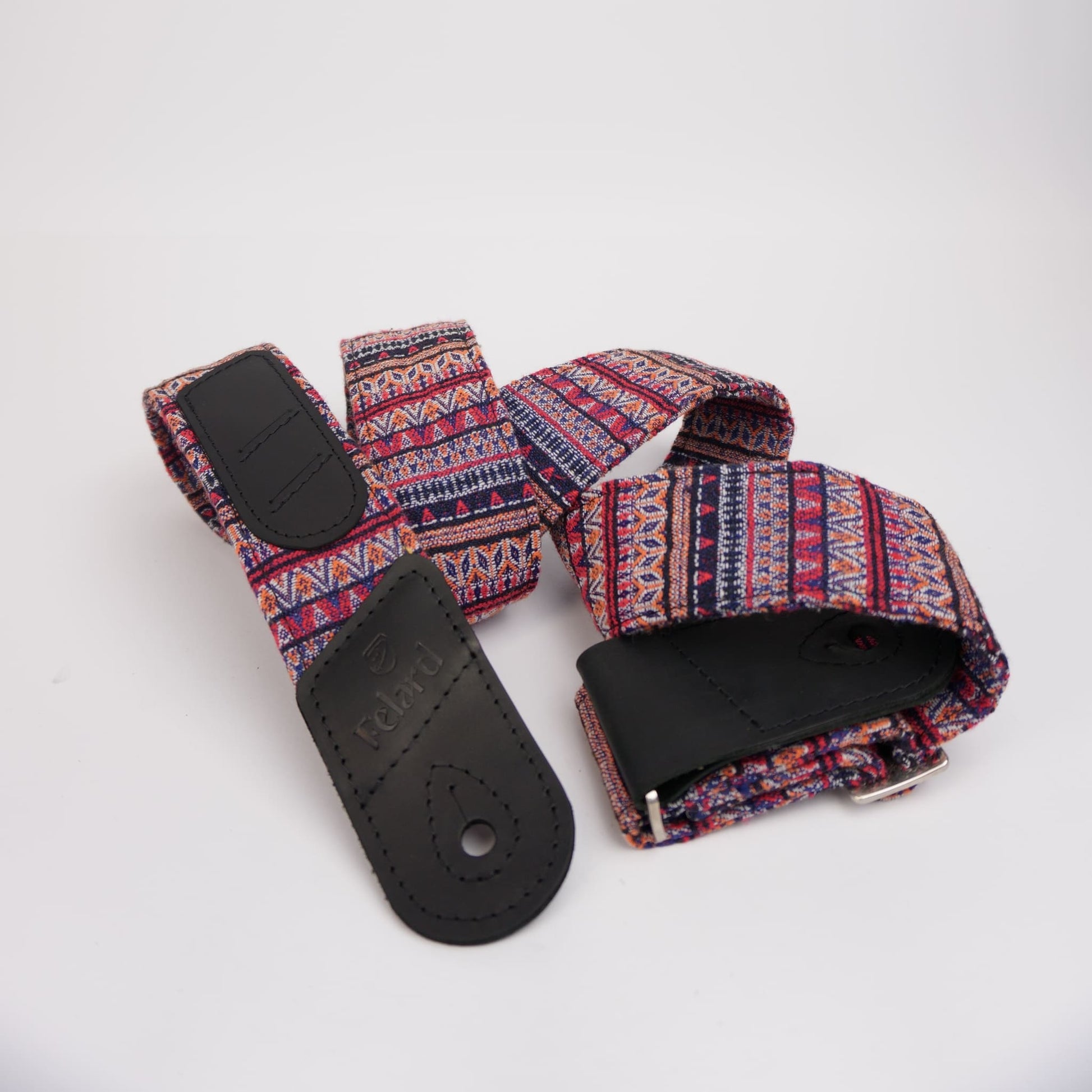 A fabric guitar strap with a colorful geometric pattern in red, blue, and white, featuring black leather end pieces and a silver metal buckle, displayed on a white background.