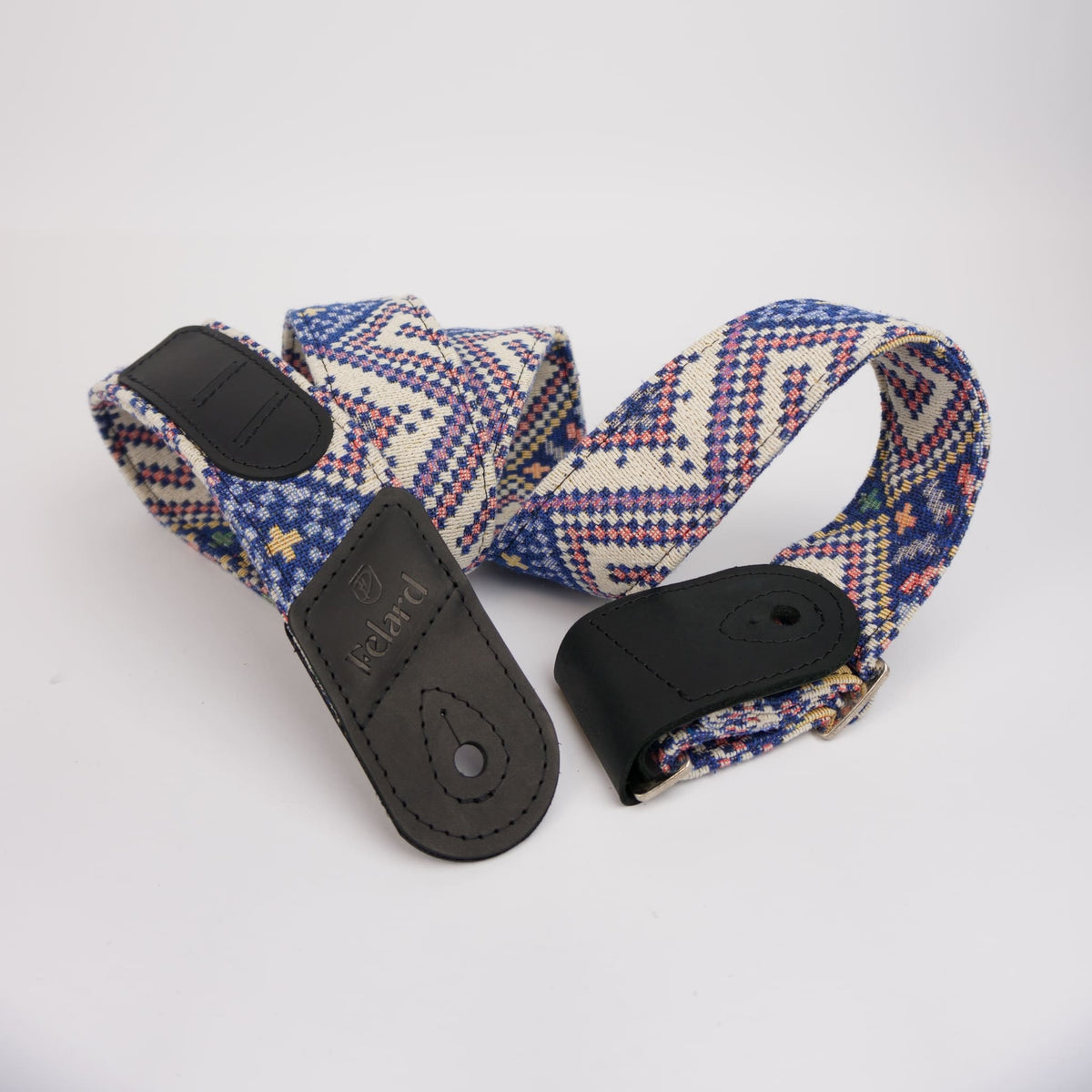 A guitar strap with a blue, red, and cream geometric patterned fabric body and black leather ends, one of which is embossed with the brand name 'Felard'.