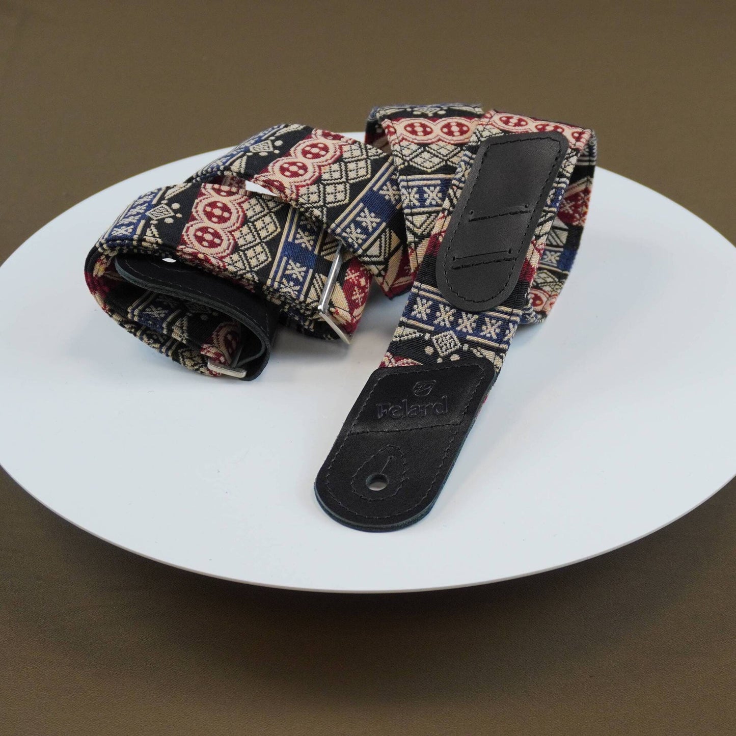 guitar strap with a Southwestern geometric pattern in red, blue, beige, and black, featuring a black leather end piece with a thumbhole, displayed on a white plate against a brown background.