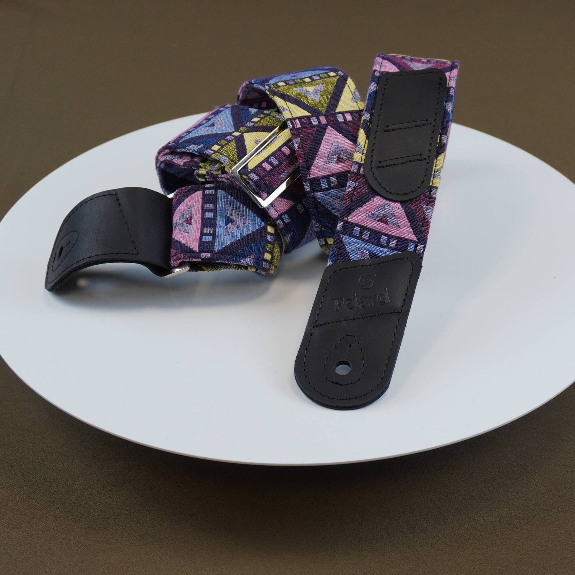 Guitar strap with a colorful geometric pattern in purple, pink, yellow, and blue, featuring black leather end caps, displayed on a white plate against a brown background.