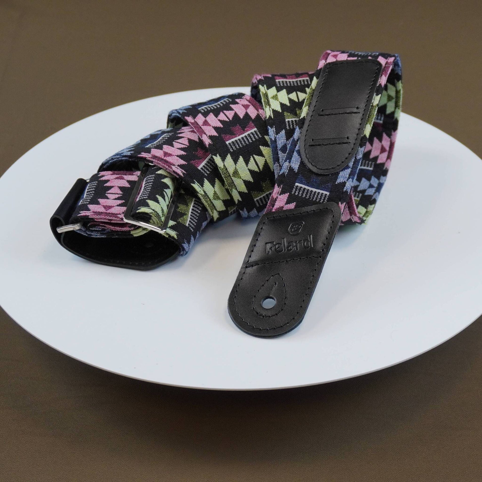 Guitar strap with black leather end caps resting on a white plate against a brown background. The fabric features a geometric triangle pattern in pink, green, and white on a black base.