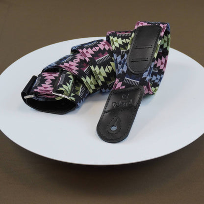 Guitar strap with black leather end caps resting on a white plate against a brown background. The fabric features a geometric triangle pattern in pink, green, and white on a black base.