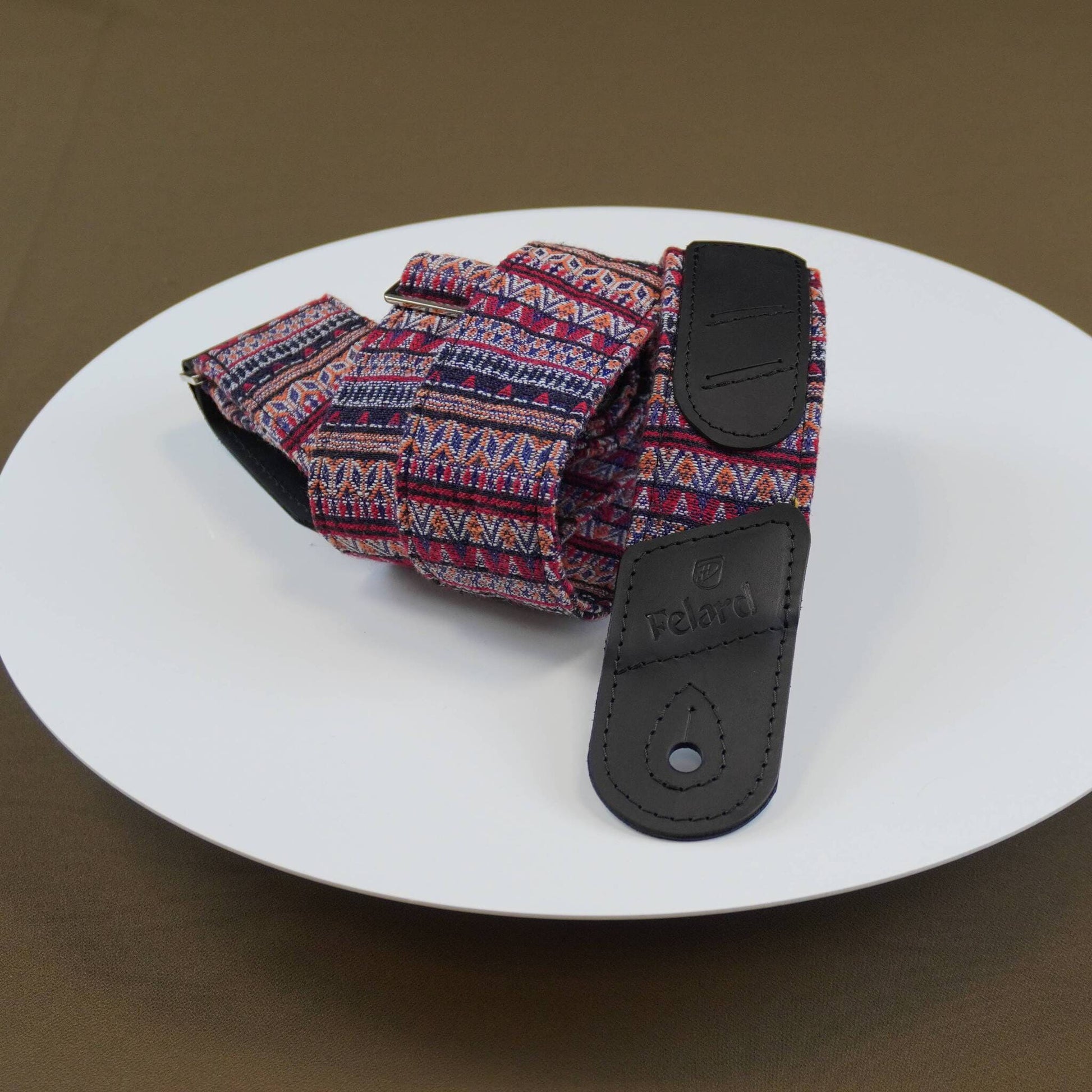 A patterned guitar strap with a multicolor geometric design and black leather end pieces, displayed on a white plate against a brown background.