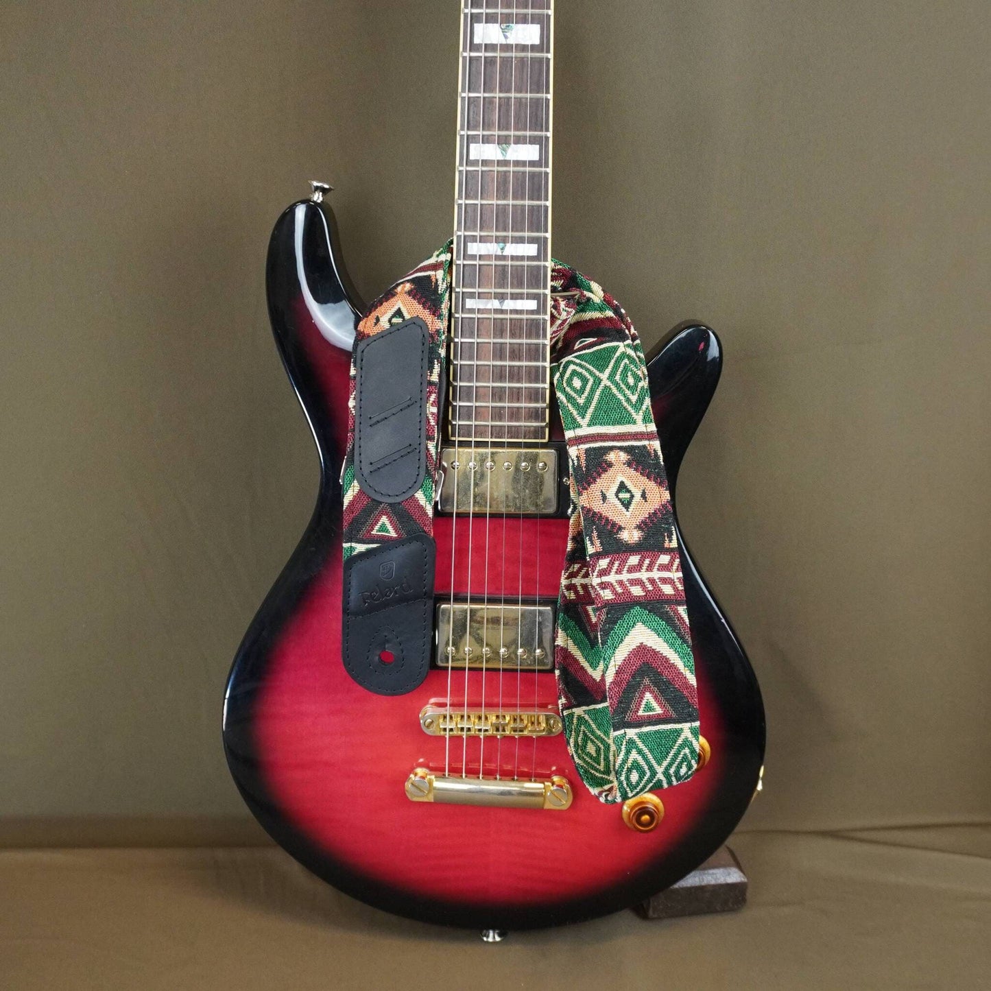 a colorful geometric patterned strap and black pickguard, displayed on a stand against a plain brown background.