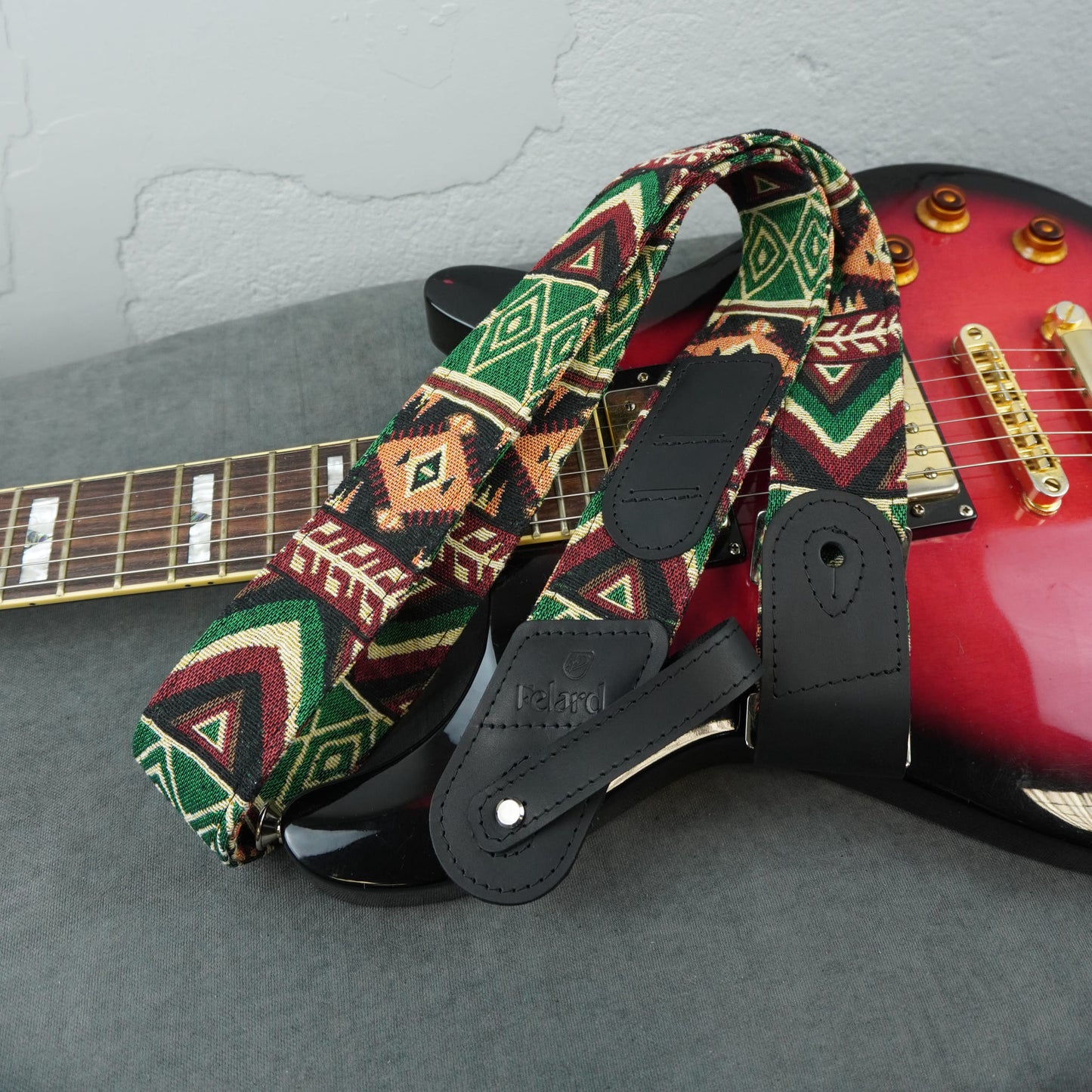 A guitar strap with a Southwestern geometric pattern in green, red, black, and beige, shown draped over a red electric guitar on a gray surface.
