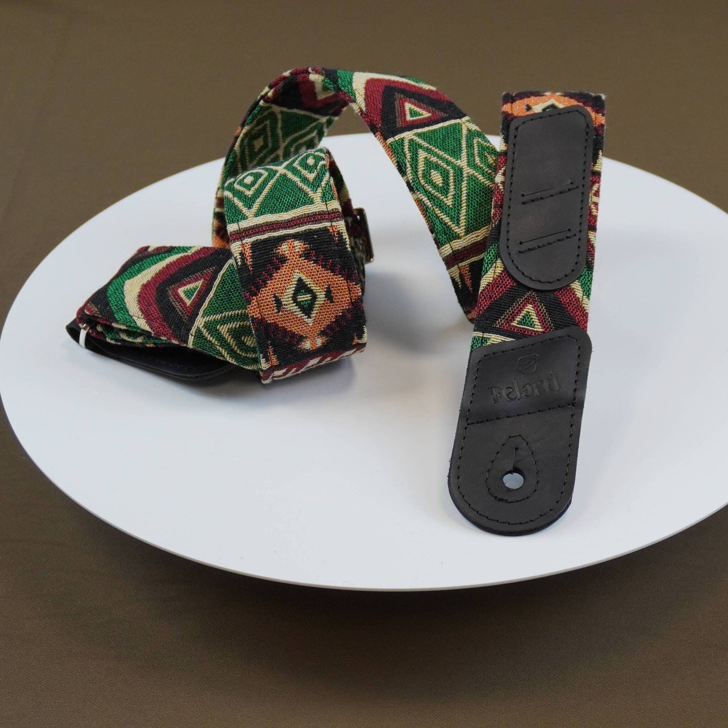 A patterned guitar strap with a geometric Southwestern design in green, red, and brown, featuring black leather end pieces, displayed on a white plate.