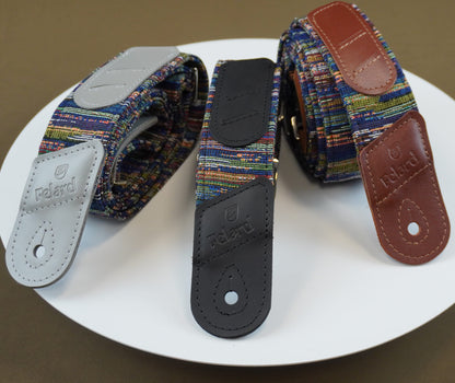 Three guitar or ukulele straps with multicolor blue rain fabric cores and leather end pieces in gray, black, and brown, arranged on a white plate against a brown background.
