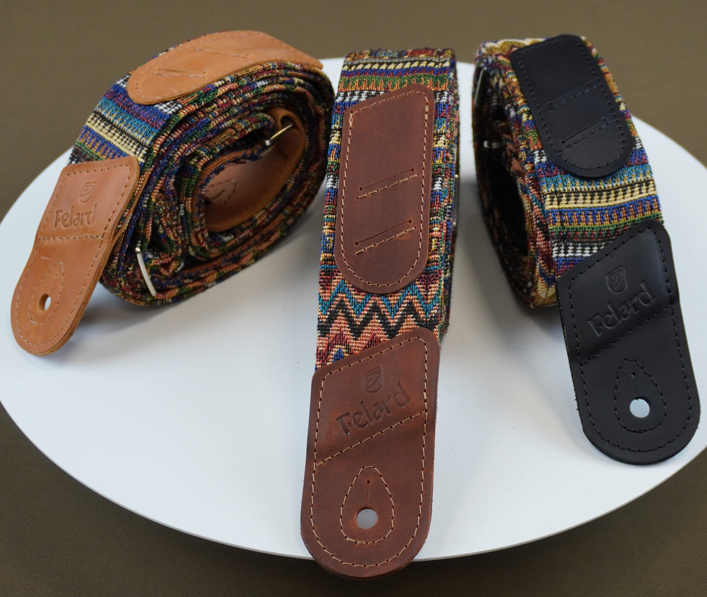 guitar straps from the brand Redbud are displayed on a white circular plate against a dark brown background. One strap is coiled, showing its multicolor woven pattern and brown leather end piece.