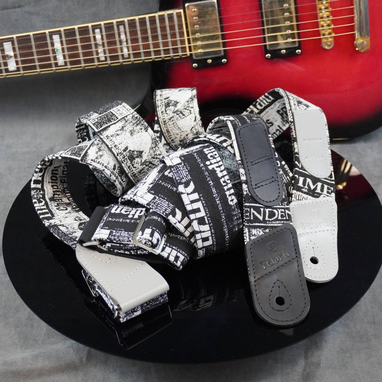 Three black and white newspaper print guitar straps with black leather end pieces are displayed on a black reflective plate, with a red electric guitar blurred in the background.