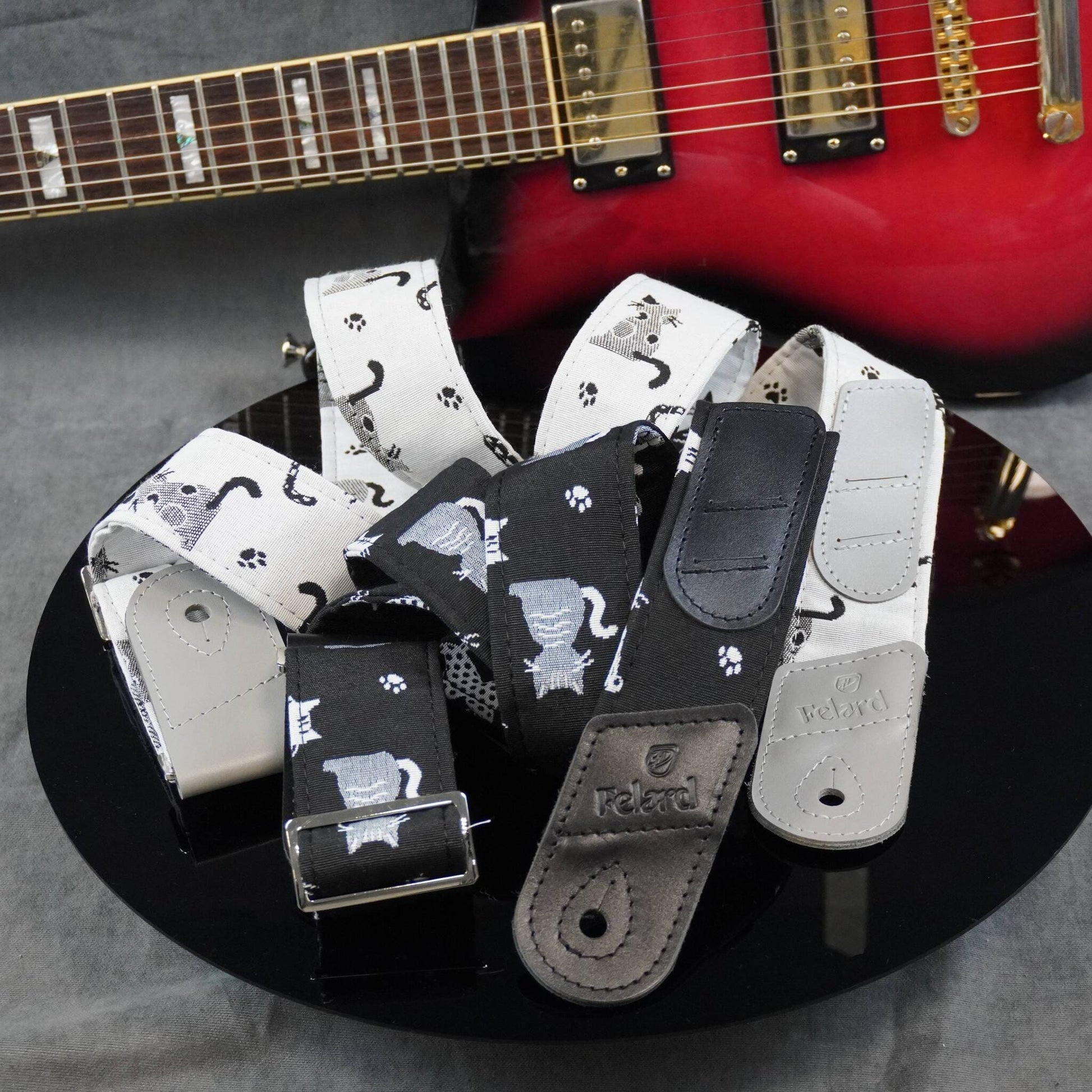 A set of guitar shoulder straps with black and white cat-themed patterns, displayed on a black reflective surface with a red electric guitar in the background.