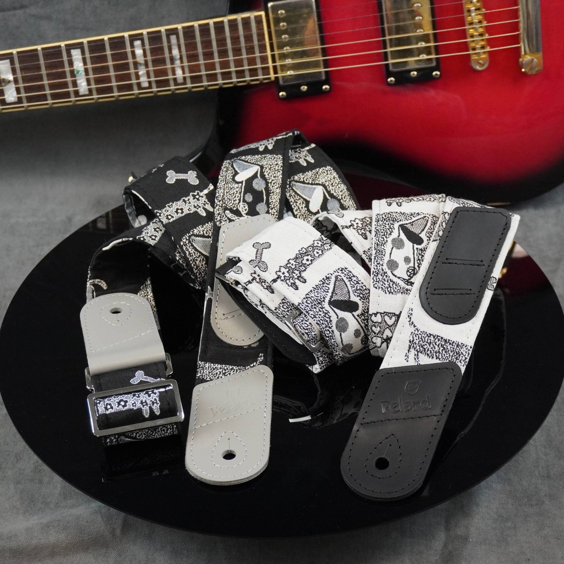 A set of guitar strap covers with a black and white pattern, displayed on a black reflective surface with a red electric guitar in the background.