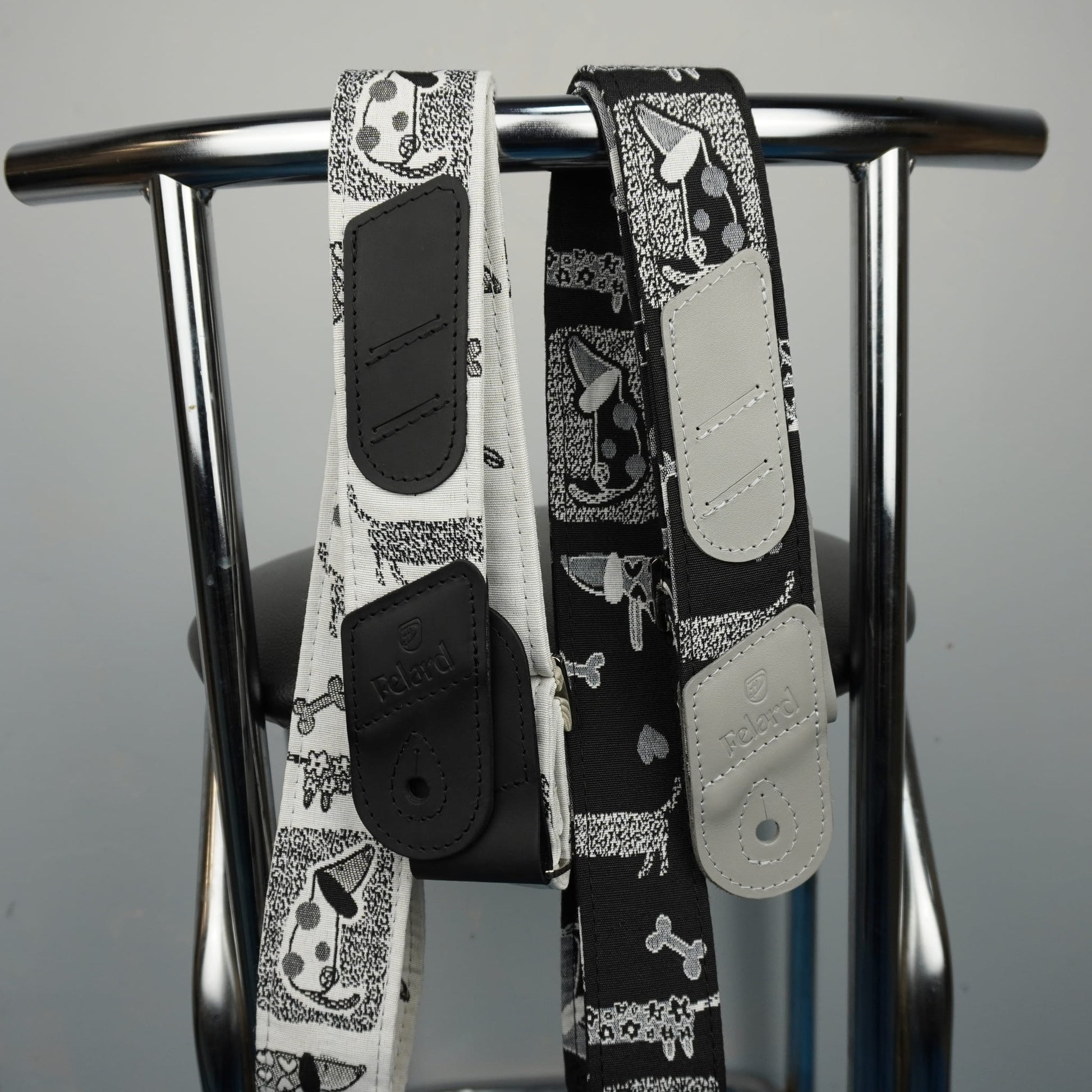 Two dog-print leather shoulder straps hanging from a metal stand against a plain light gray background.