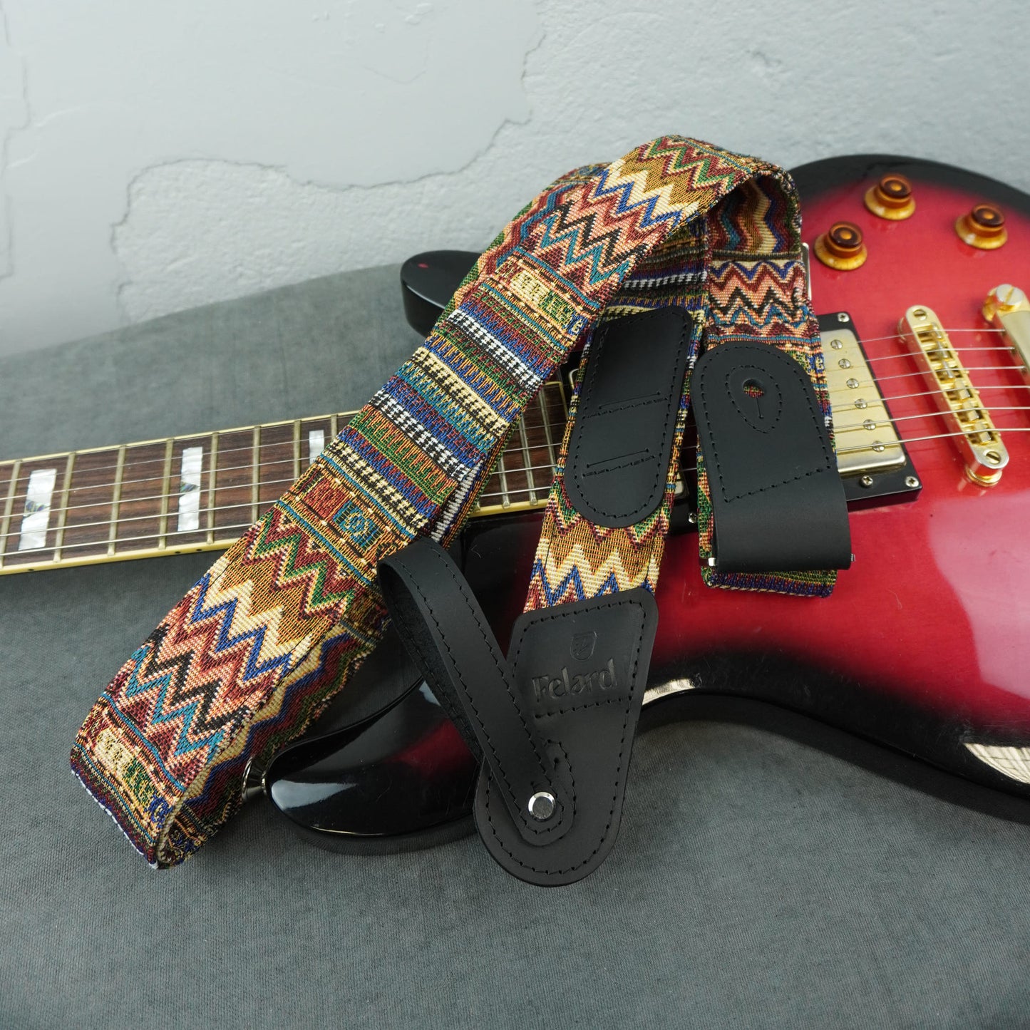 A colorful woven electric guitar strap with a geometric zigzag pattern in red, blue, yellow, and brown, featuring black leather accents, draped over a red and black electric guitar.
