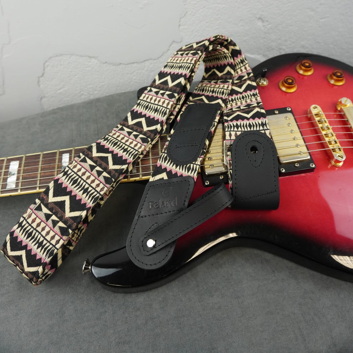 guitar strap with a black, beige, and pink geometric pattern and black faux-leather padding, draped over a red electric guitar on a gray surface.