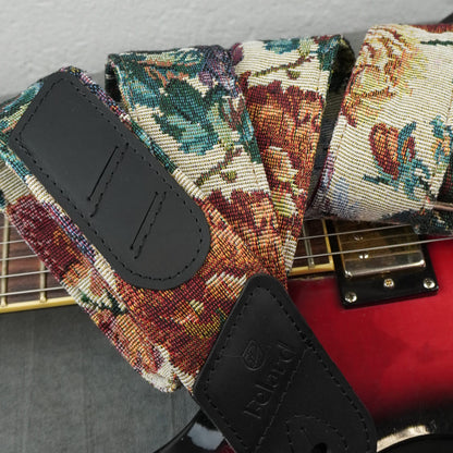 A close-up of a guitar strap with a floral needlepoint pattern in red, green, and beige, featuring black leather patches. The strap is shown draped over a red electric guitar.