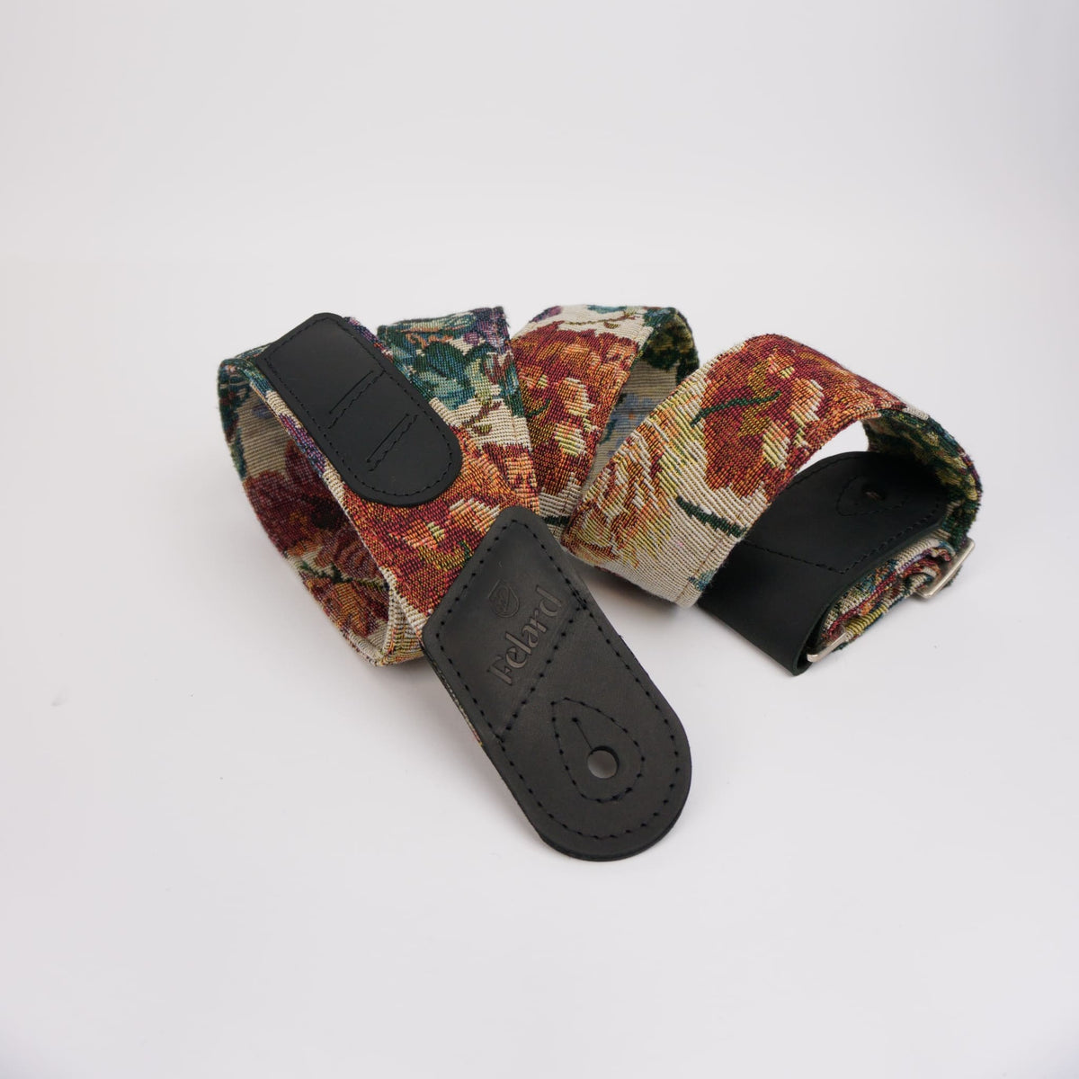 A guitar strap featuring a dense floral tapestry pattern in warm tones of red, orange, and green on a beige background, with black leather end caps and a buckle.