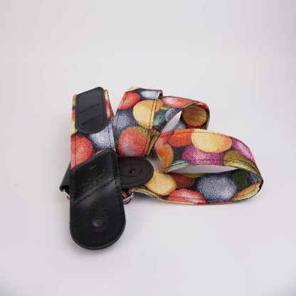A camera strap featuring a colorful print with red, yellow, orange, and green motifs on a white background, finished with black leather hardware.