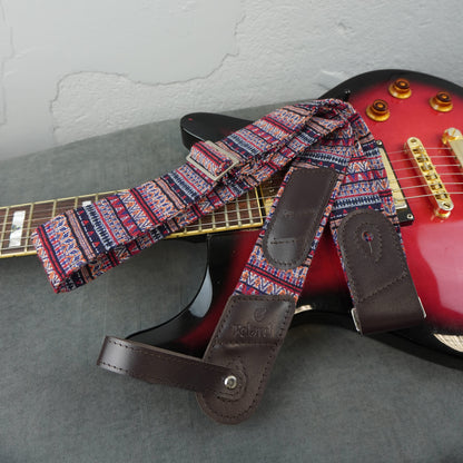 guitar strap with a red, blue, and white geometric pattern and dark brown leather ends, laid across a red electric guitar on a gray surface.