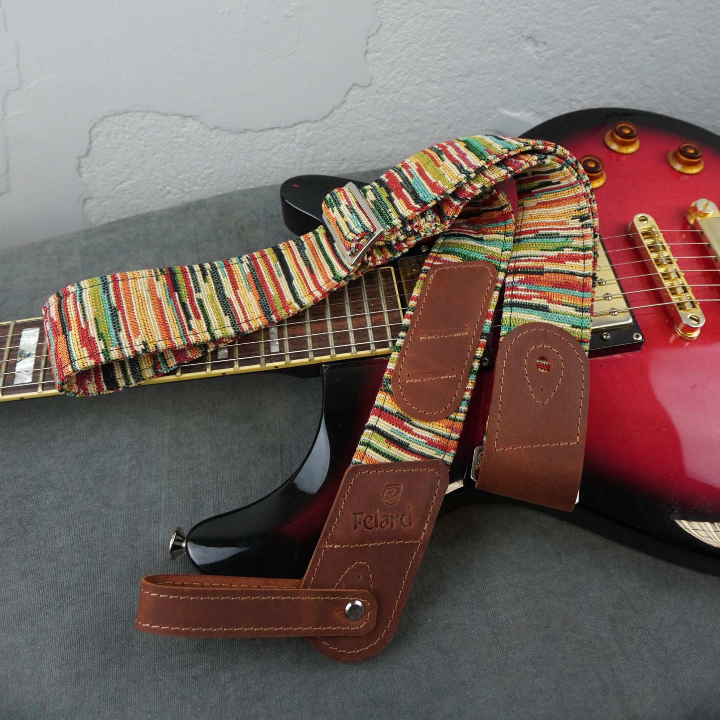 guitar strap with a colorful striped woven pattern and brown leather end pieces, draped over a red electric guitar on a gray surface.