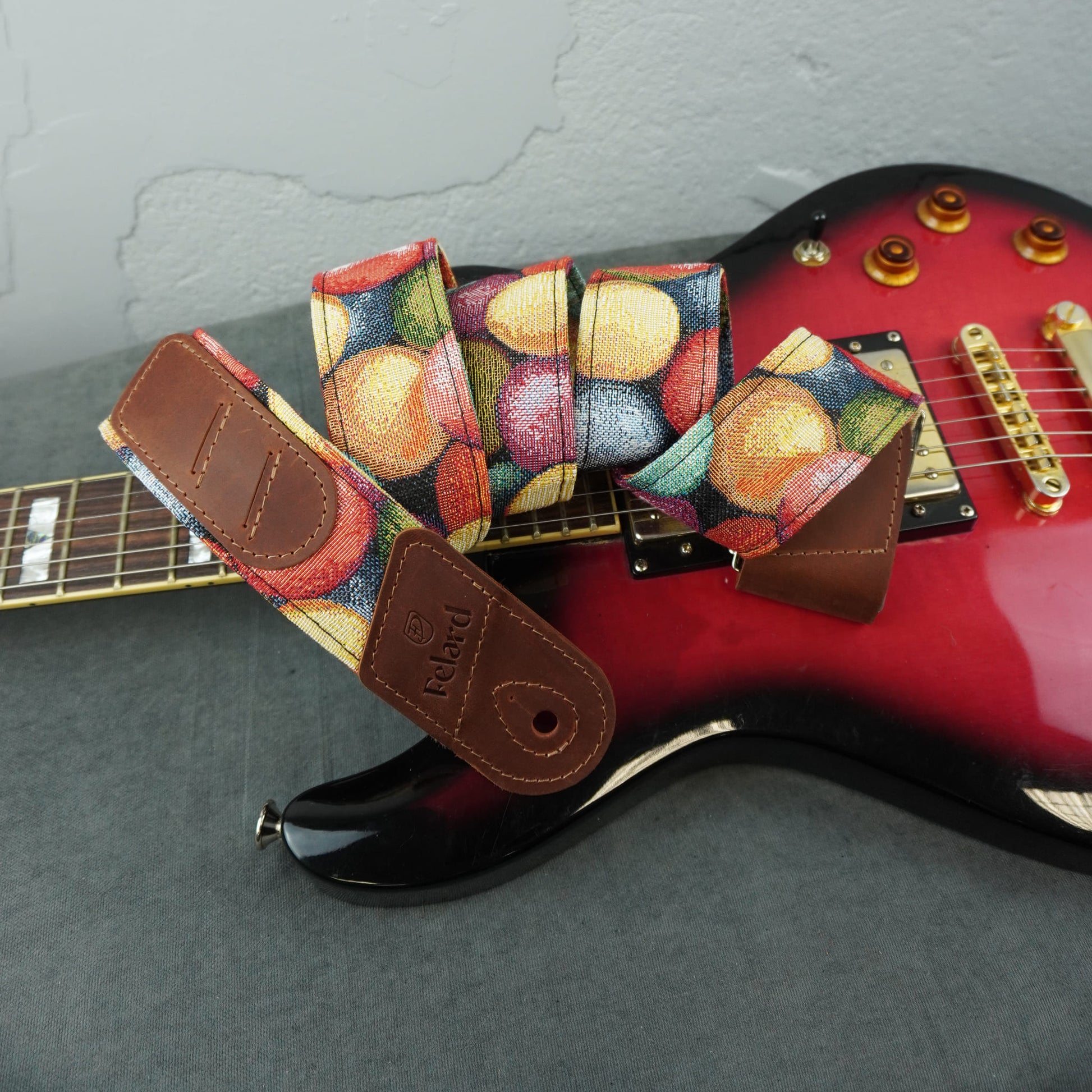 A guitar strap with a colorful fruit print and brown leather ends, draped over a red electric guitar on a gray surface.