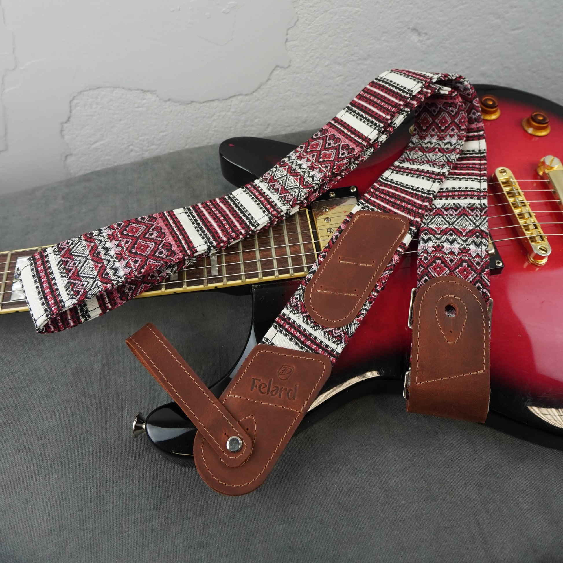 guitar strap with a red, white, and black geometric pattern and brown leather ends, draped over a red electric guitar on a gray surface.