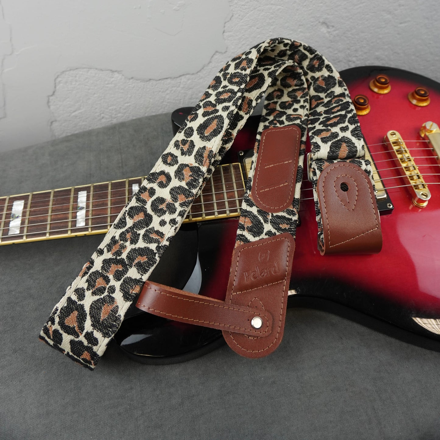 A leopard print guitar strap with brown leather accents, draped over a red electric guitar on a gray surface against a light gray wall.